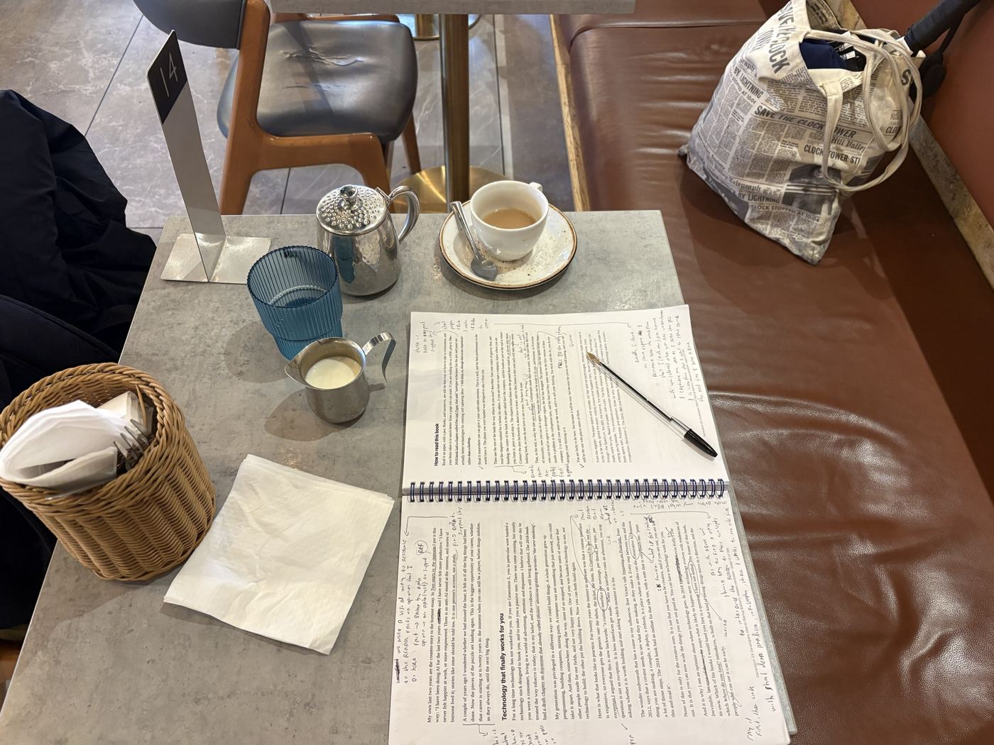 The printed release candidate open at pages 12 and 13 on a café table, with a pen, a teapot, a milk jug, a cup of tea, and a tote bag reading Save the Clock Tower on the seat